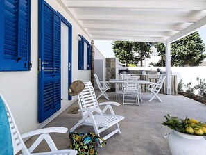 A covered outdoor seating area with white chairs and a table, blue shutters, and a white pot with lemons. - Belvilla by OYO Spuma di Mare - Riccio (San Vito Lo Capo)