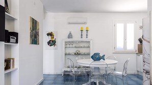 A dining area with a round table and chairs, a white cabinet with glassware, and a blue tiled floor. - Belvilla by OYO Spuma di Mare - Riccio (San Vito Lo Capo)