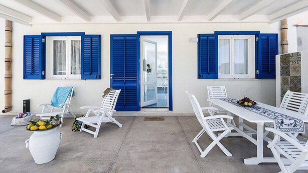 A covered outdoor area with white furniture, blue shutters, and a glass door leading to a pool. - Belvilla by OYO Spuma di Mare - Riccio (San Vito Lo Capo)