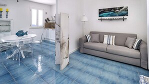A living room with a grey sofa, a dining table with chairs, and a kitchen area with white cabinets. - Belvilla by OYO Spuma di Mare - Riccio (San Vito Lo Capo)