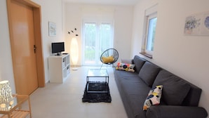 Un salon moderne avec un canapé gris, un fauteuil noir, un téléviseur posé sur un meuble blanc et une grande fenêtre.