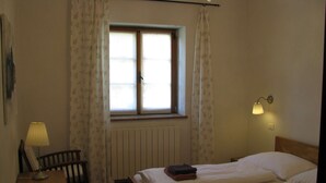 Habitación de hotel con dos camas, un radiador, una ventana con cortinas, un escritorio con lámpara y una lámpara de pared.