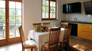 Un comedor con una mesa dispuesta para dos personas, sillas de madera, mantel blanco y un centro de mesa floral. Se observan dos ventanas con marcos de madera, una zona de cocina con armarios amarillos y una campana extractora negra.