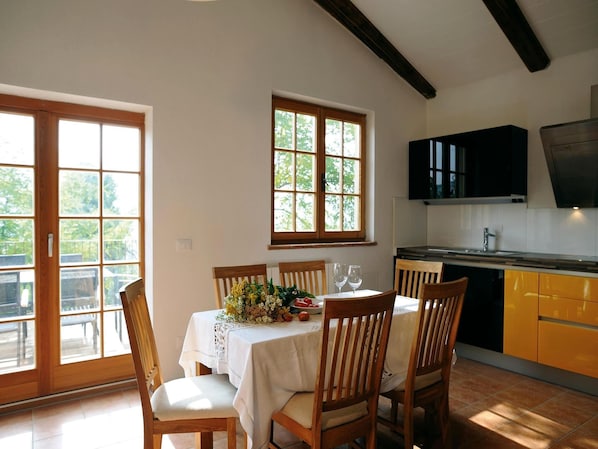 Ein Essbereich mit einem Tisch für zwei Personen, Holzstühle, einer weißen Tischdecke und einem floralen Mittelstück. Zwei Fenster mit Holzrahmen, ein Küchenbereich mit gelben Schränken und einer schwarzen Dunstabzugshaube sind zu sehen.