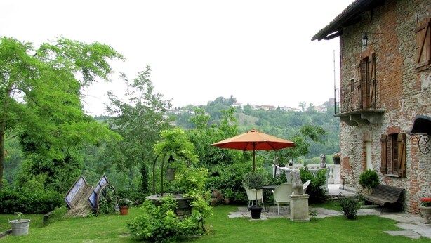 A courtyard with a grassy lawn, a red umbrella, outdoor furniture, and a stone building with a balcony. - Mansion in Langhe With Pool and Winery Charm (Bastia Mondovì)