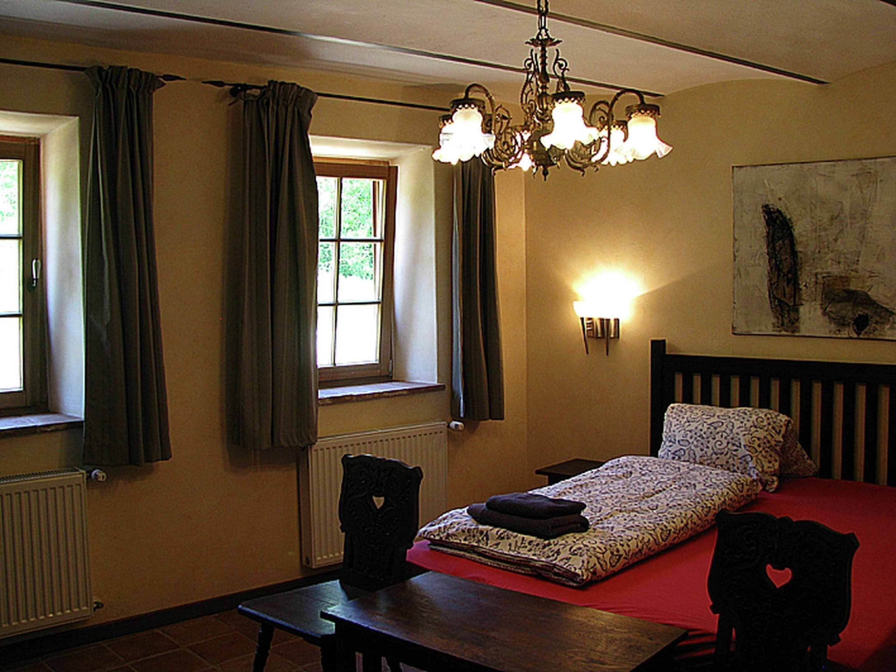 A bedroom with a bed, a table, a chair, a radiator, a window with curtains, and a wall-mounted light fixture.