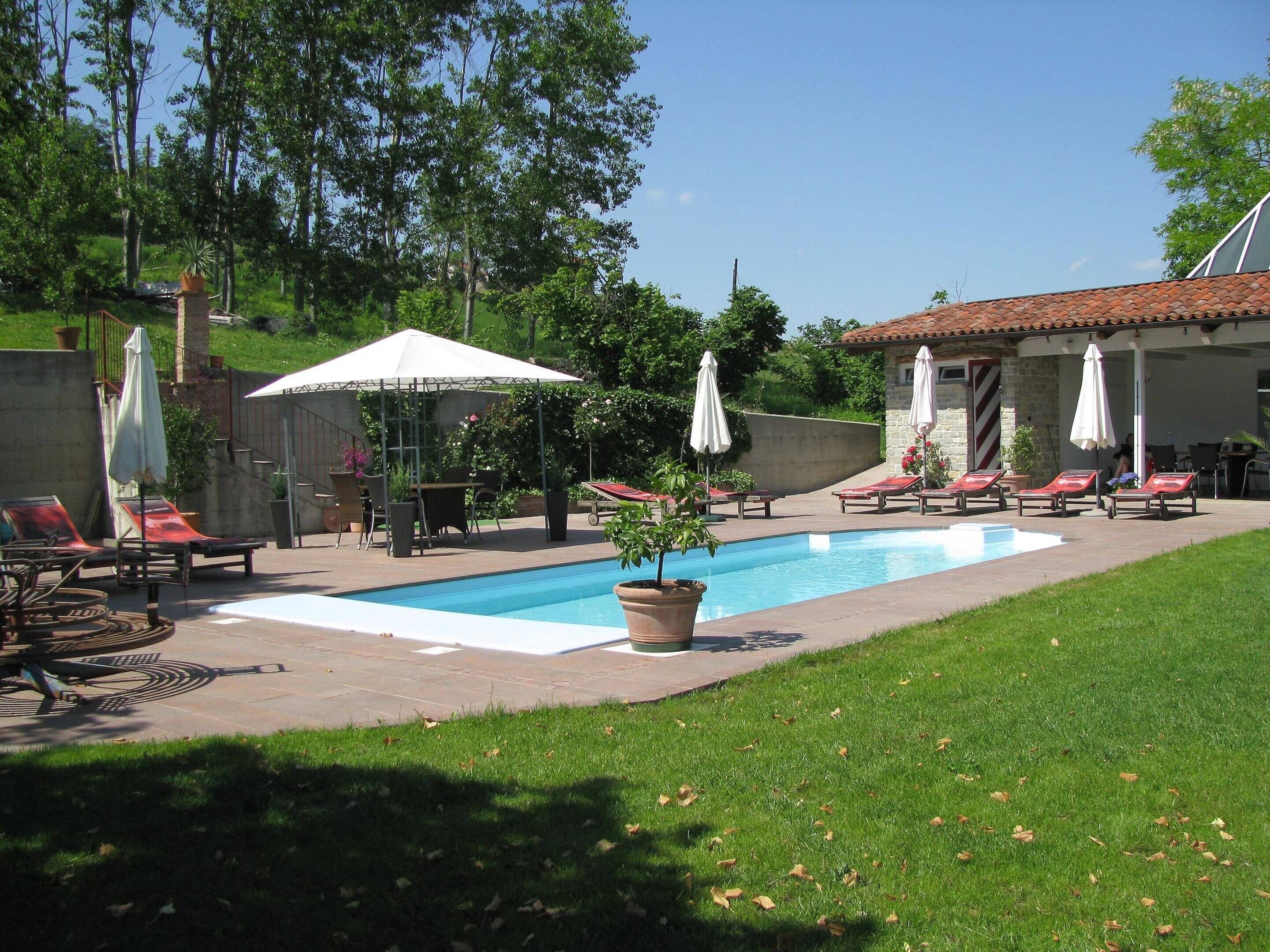 A swimming pool with lounge chairs and umbrellas, surrounded by greenery and a stone building.