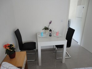 Une petite pièce propre, avec une table blanche et deux chaises noires. On y trouve, à gauche, une plante en pot, une petite table en bois sur laquelle est posé un vase de fleurs, et un mur blanc avec une porte et un interrupteur.