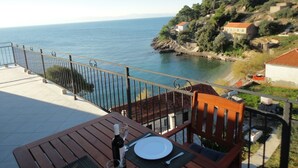 Una terrazza con un tavolo apparecchiato per due, affacciata su una zona costiera con case e una vista limpida sul mare.