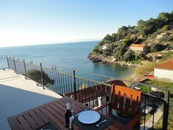 Une terrasse avec une table dressée pour deux personnes, donnant sur une zone côtière avec des maisons et offrant une vue dégagée sur la mer.