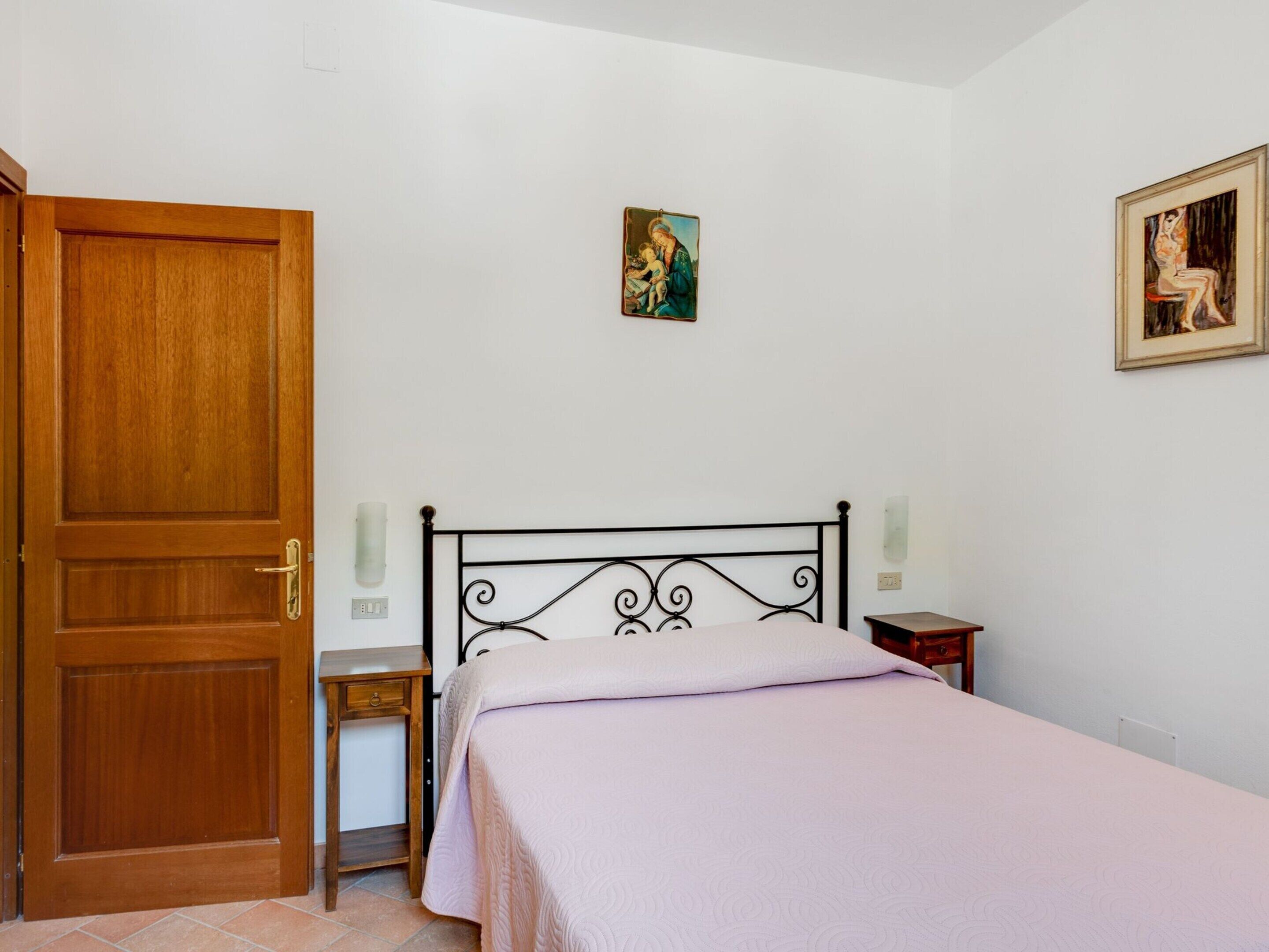 A bedroom with a wooden door, a metal bed frame, a bedside table, a nightstand, and a wall-mounted picture.