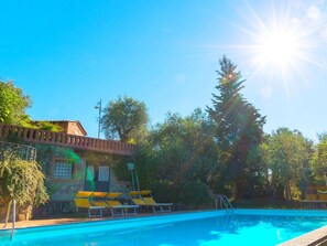A swimming pool with sunlit surroundings, a brick building with a tiled roof, and a row of yellow lounge chairs.
