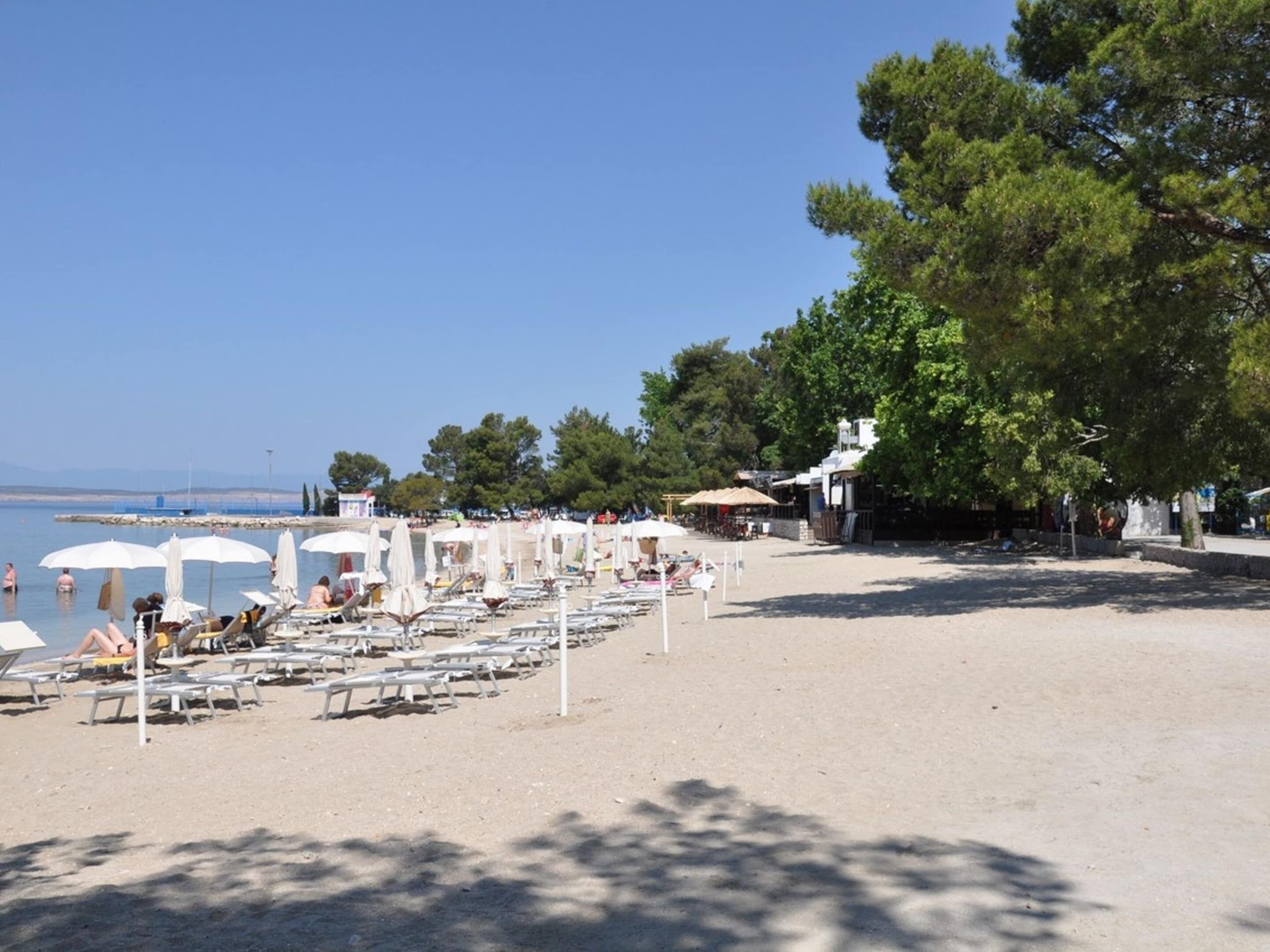 Ein Strand mit Liegestühlen und Sonnenschirmen, einem klaren Himmel und Bäumen im Hintergrund.
