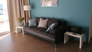 A modern living room with a dark brown sofa, a framed picture on the wall, a small white side table, a lamp, and a TV on a stand. - Apartment by the Beach in Dalmatia (Bilice)