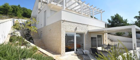 A modern two-story house with a stone facade, a balcony with a white railing, and a covered outdoor seating area with lounge chairs.