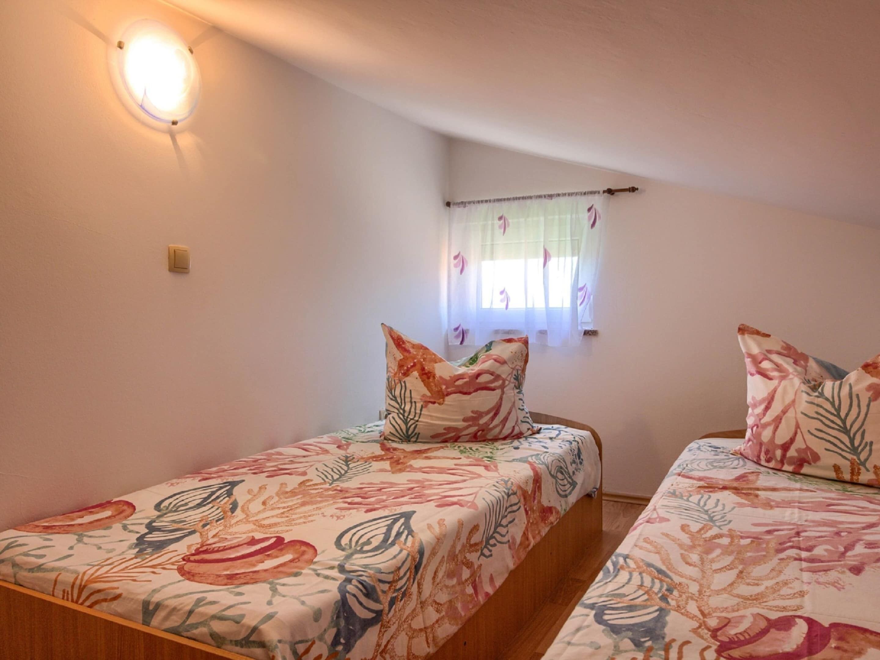 A small, cozy room with two single beds, each with floral-patterned bedding. There is a window with sheer curtains and a wall-mounted light fixture.