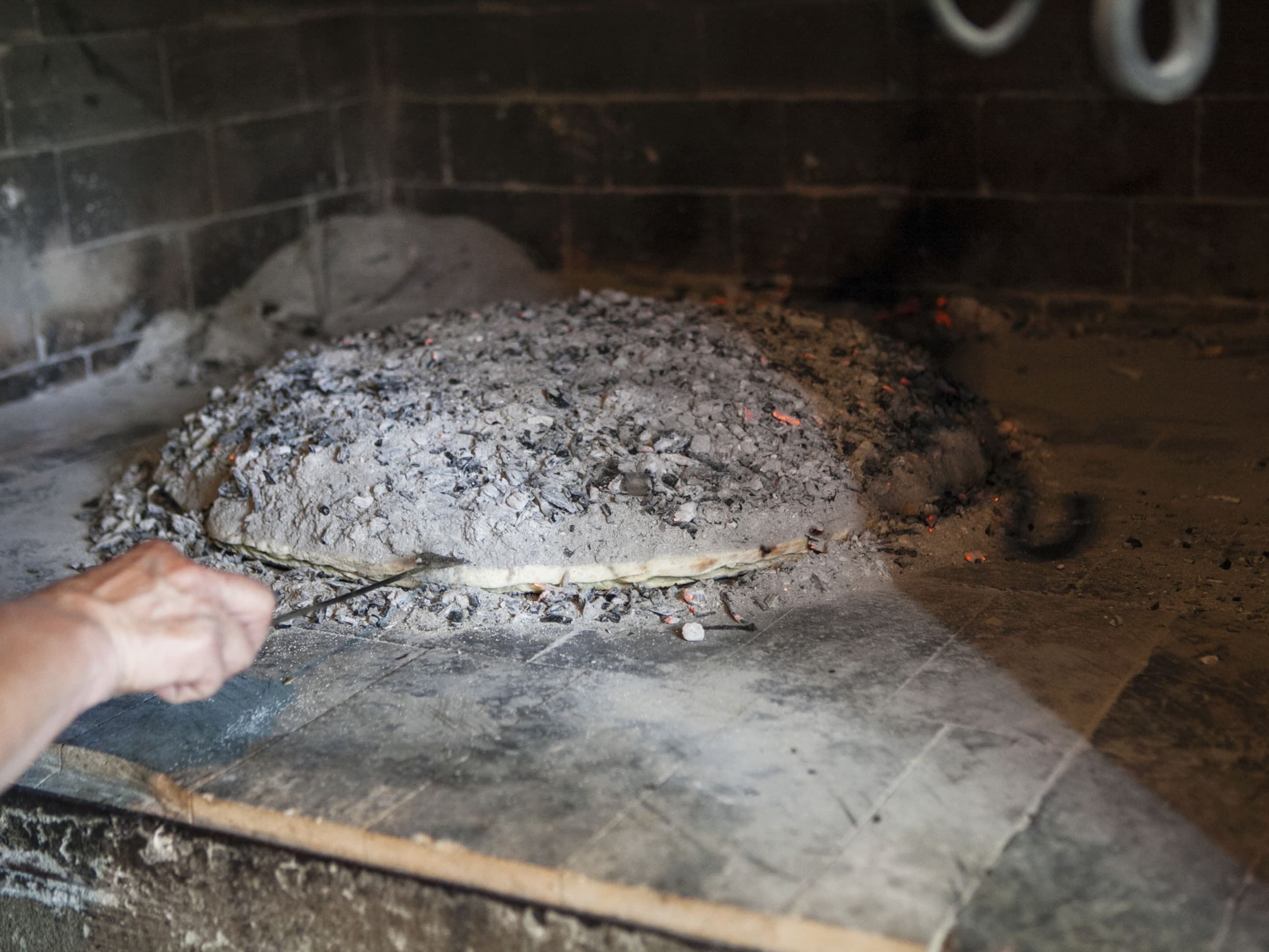 Une grande pizza cuite dans un four en briques, et une personne qui utilise un ustensile pour la manipuler.
