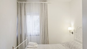 Een slaapkamer met een bed, een raam met dunne gordijnen, een bedlamp en een opgevouwen handdoek op het bed.