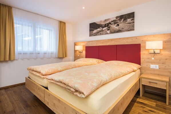 A double bed with a red headboard, two bedside tables with lamps, a wooden floor, and a window with sheer curtains.