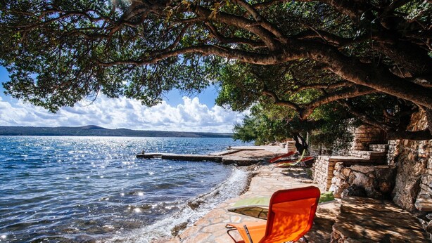 Un chemin de pierre mène à un espace de détente aménagé au bord du lac, avec une chaise rouge et une table, d’où l’on a une vue sur l’eau.
