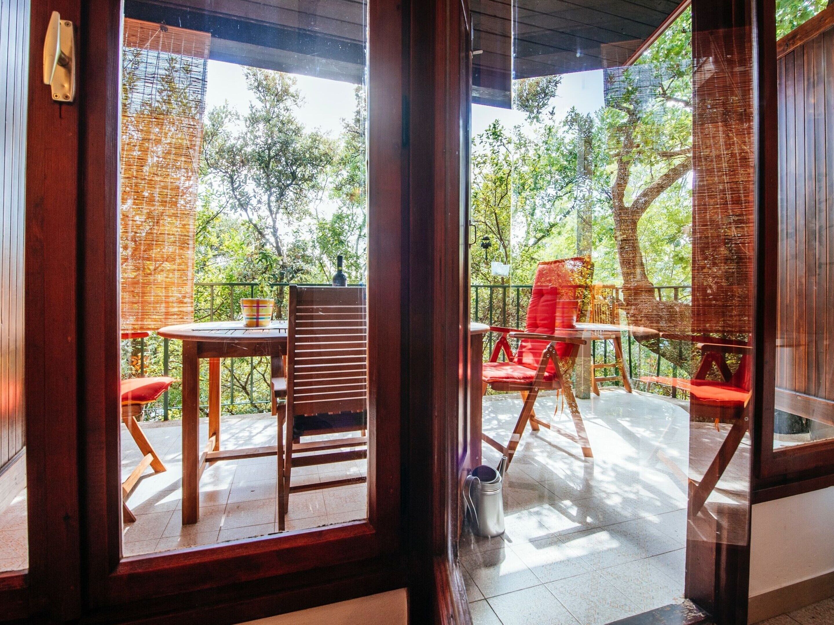 An outdoor patio with a wooden table and chairs, a metal bucket, and a view of trees through a wooden fence.