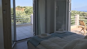 A bedroom with two beds, a balcony with a view of greenery and the sea, and a chair.