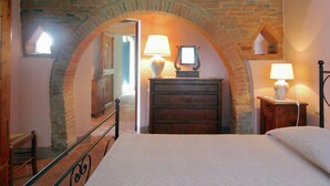 A bedroom with a stone archway, wooden dresser, two bedside lamps, and a bed with a patterned bedspread.