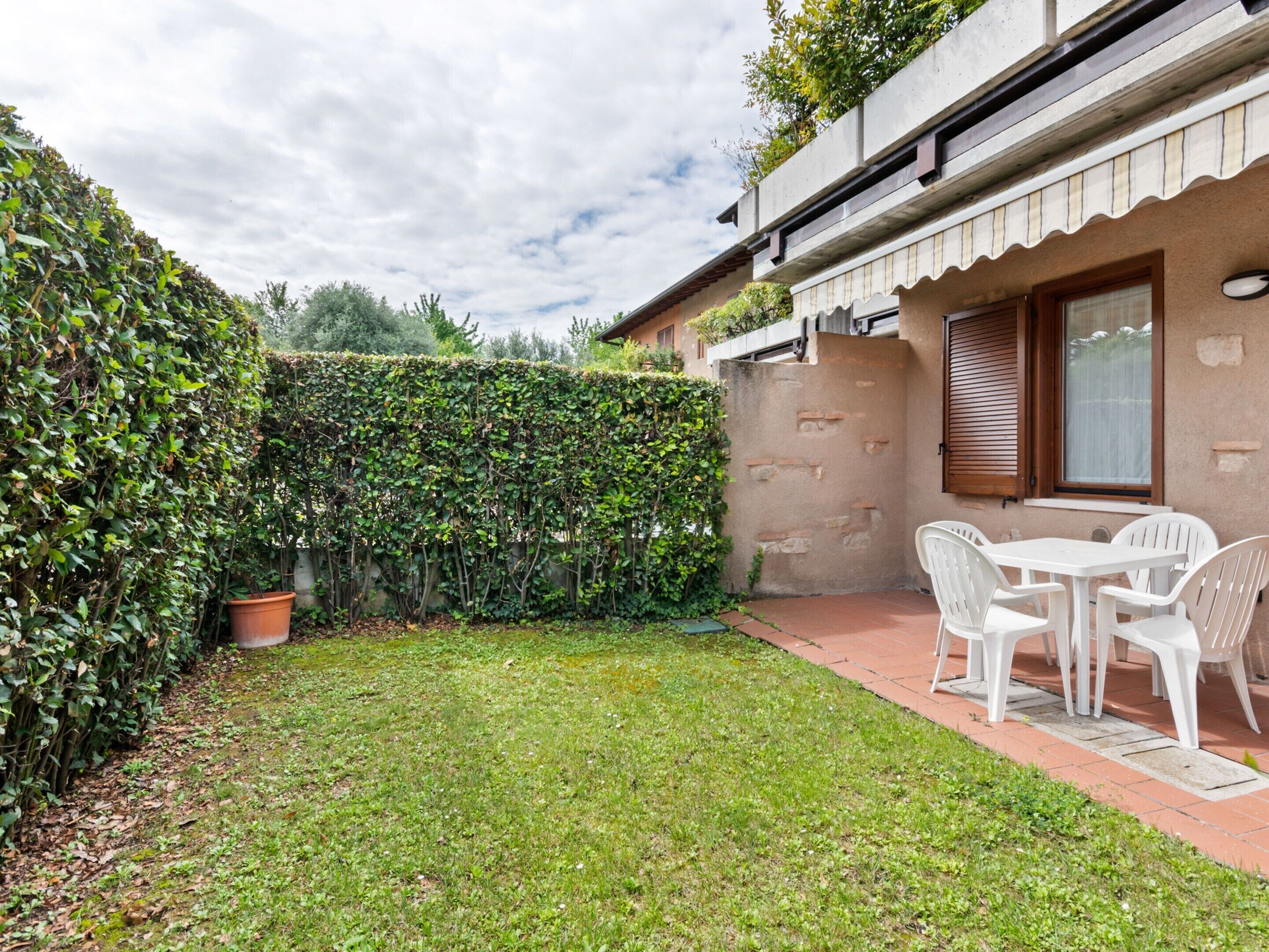 A patio with a table and chairs, a hedge, and a building with a window and a door.