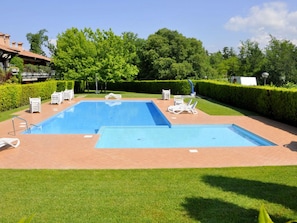 A rectangular outdoor swimming pool with two separate sections, surrounded by a brick patio. There are lounge chairs and a small table with a chair, and a hedge in the background.