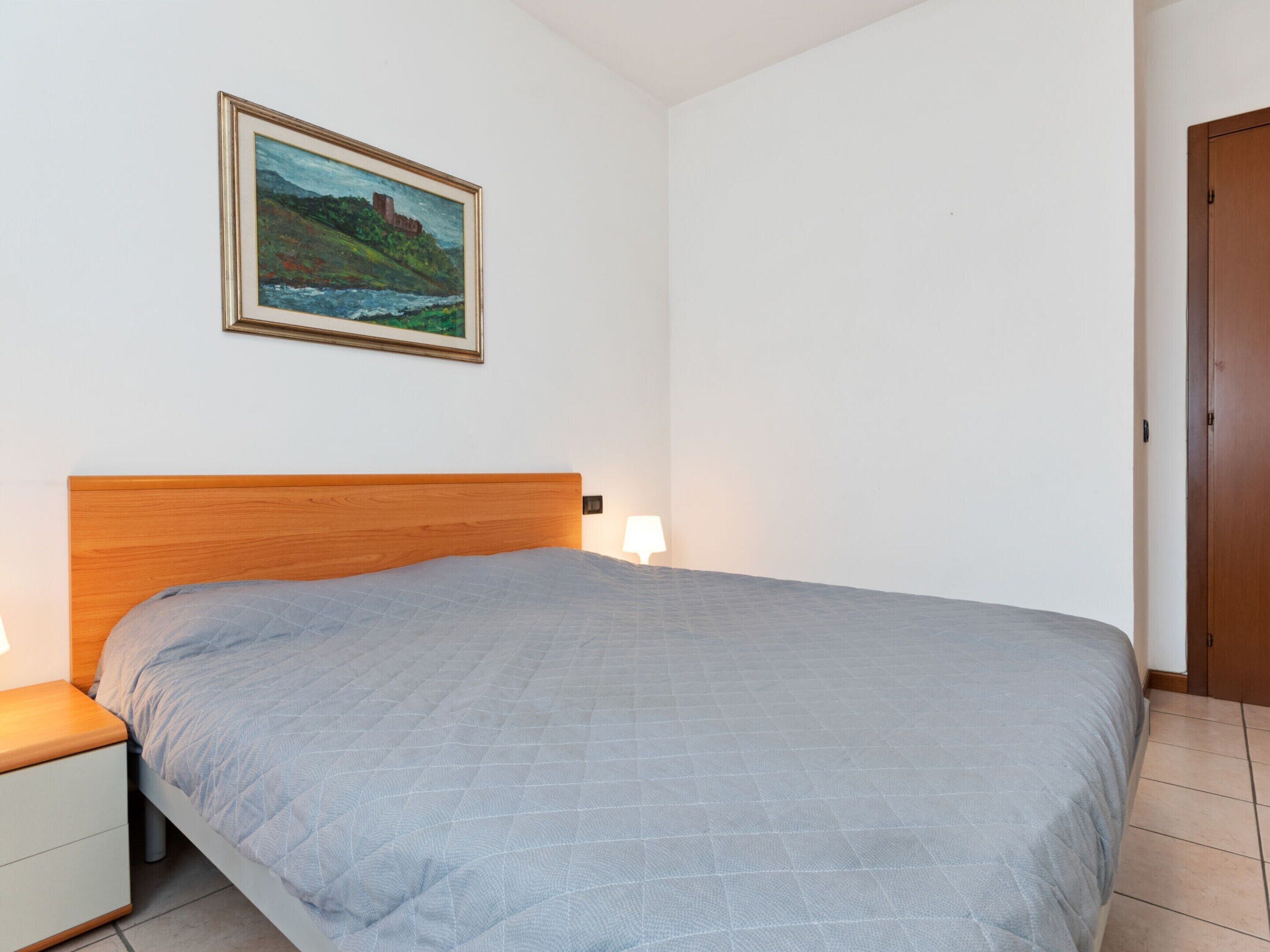 A bedroom with a wooden headboard, a single bed with a grey quilt, a bedside table with a lamp, and a framed painting on the wall.