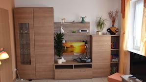 A modern living room with a flat-screen TV, wooden cabinets, a bookshelf, and a potted plant.