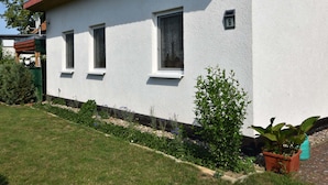 A white building with three windows and a small garden area with plants and a potted plant.