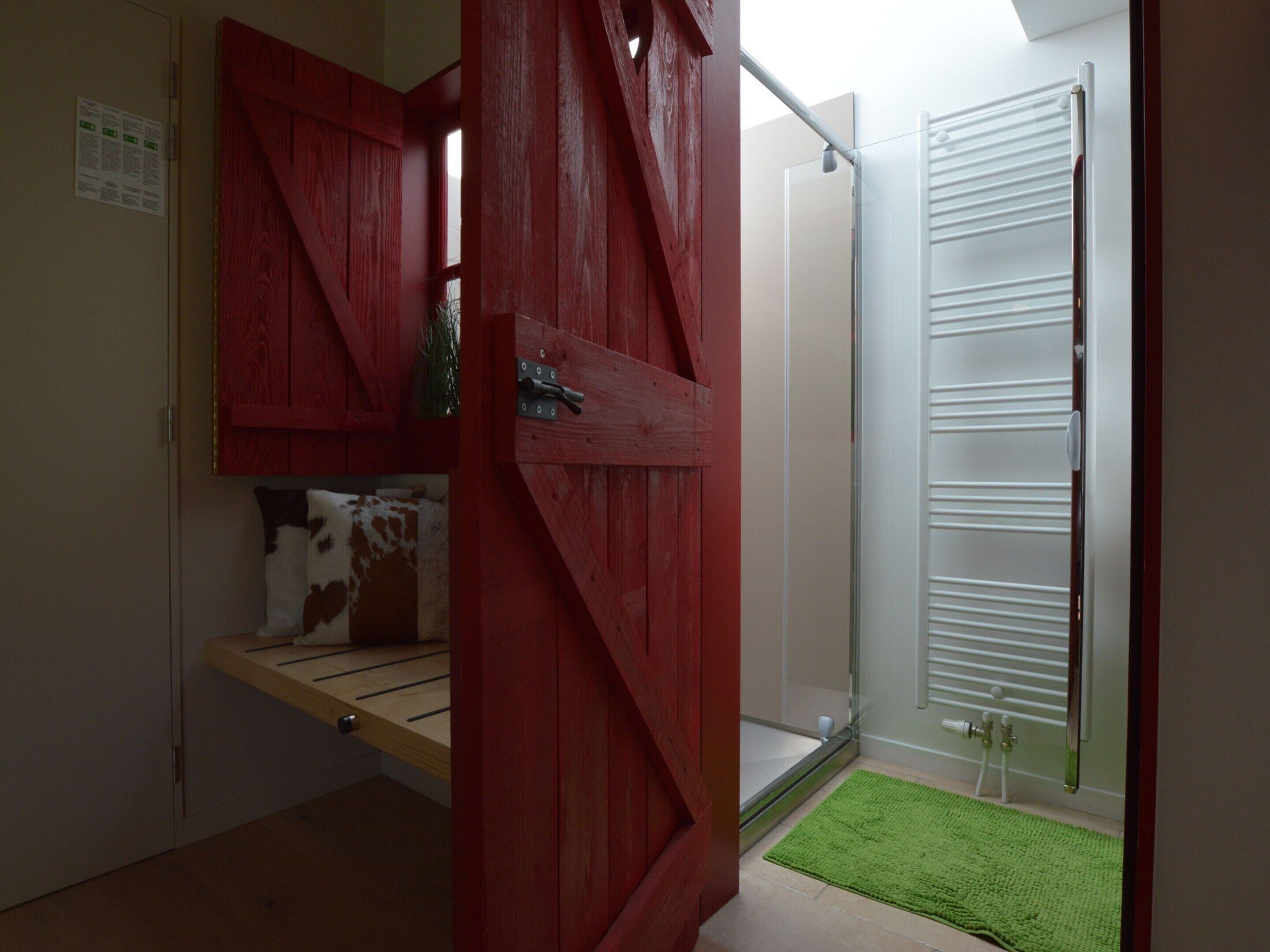 Ein Zimmer mit roter Tür, einer kleinen Sitzbank, einem grünen Teppich und einem weißen Heizkörper.