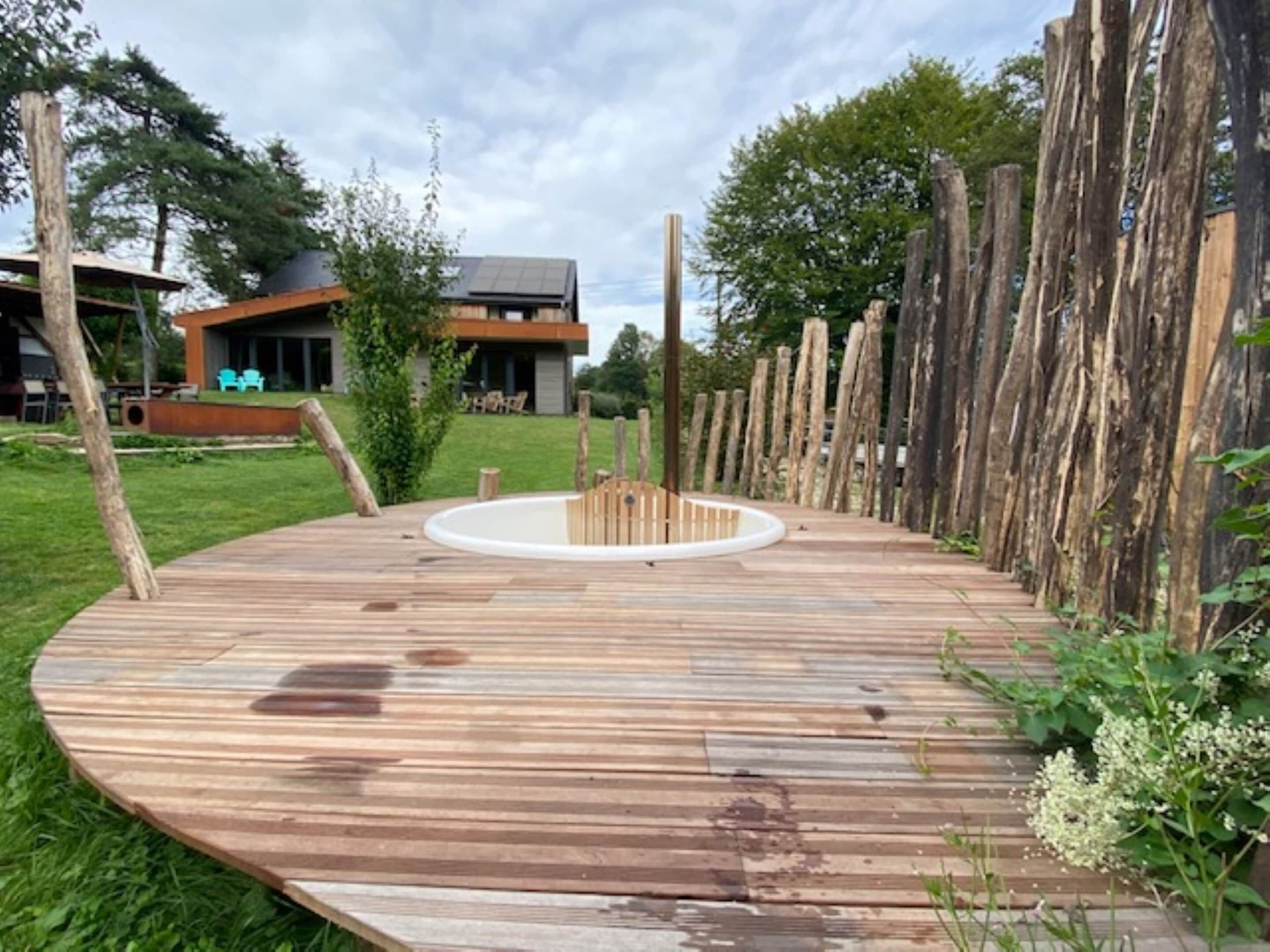 Una terraza de madera con una zona de estar circular, rodeada por un cerco de madera y follaje.