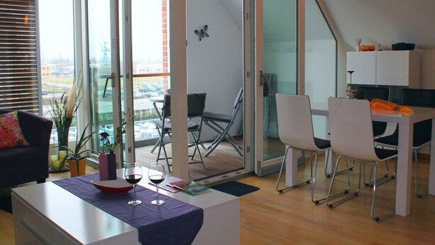 A modern living room with a glass door leading to a balcony, a dining area with a table and chairs, and a kitchen area with cabinets. - Charmante Ferienwohnung in Wismar am Hafen (Wismar)