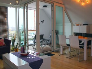 Ein modernes Wohnzimmer mit Glastür zum Balkon, einem Essbereich mit Tisch und Stühlen sowie einer Küchenzeile mit Schränken.