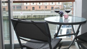 A view from a balcony with a table and two chairs, overlooking a river and a building. - Charmante Ferienwohnung in Wismar am Hafen (Wismar)