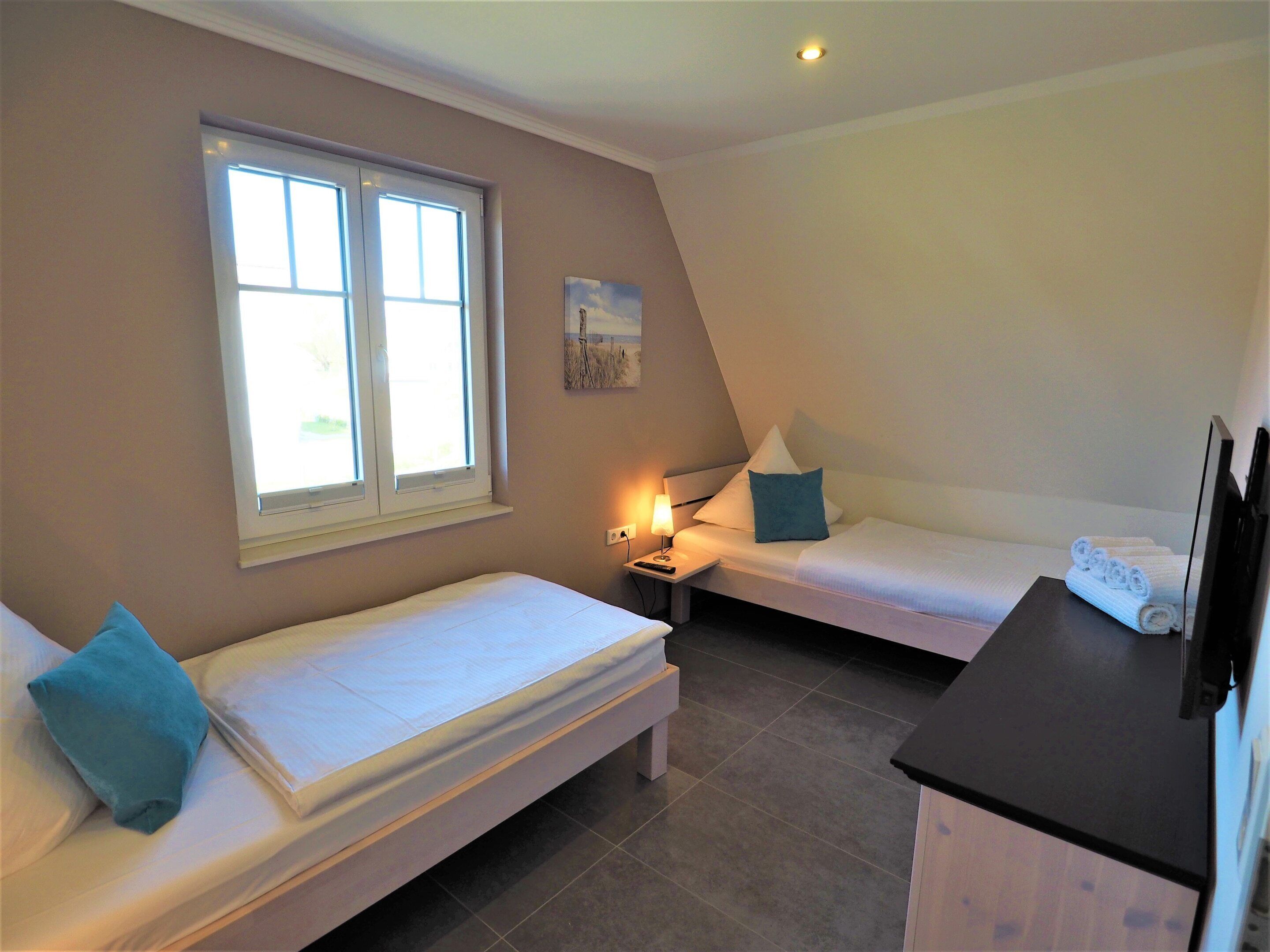 A small, well-lit room with two single beds, a nightstand with a lamp, a TV, and a window providing natural light.