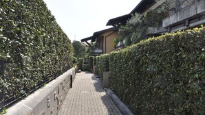 A narrow paved pathway flanked by dense hedges, leading to a building with a sloped roof and a balcony with potted plants.