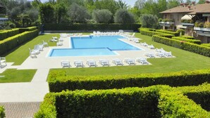 A rectangular swimming pool surrounded by white lounge chairs, with neatly trimmed hedges and a clear blue sky.