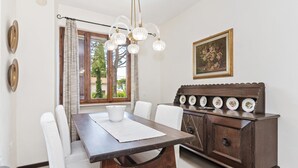A dining area with a wooden table, white chairs, a chandelier, a cabinet with plates, and a painting on the wall. - Spacious Lake Garda Villa With Private Pool, Garden and Direct Lake Access (Lazise)