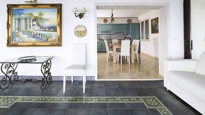 A dining area with a table and chairs, a painting, wall sconces, and a kitchen visible through an open doorway. - Exclusive Villa With Panoramic Swimming Pool and Hot Tub, Near the Sea (Letojanni)