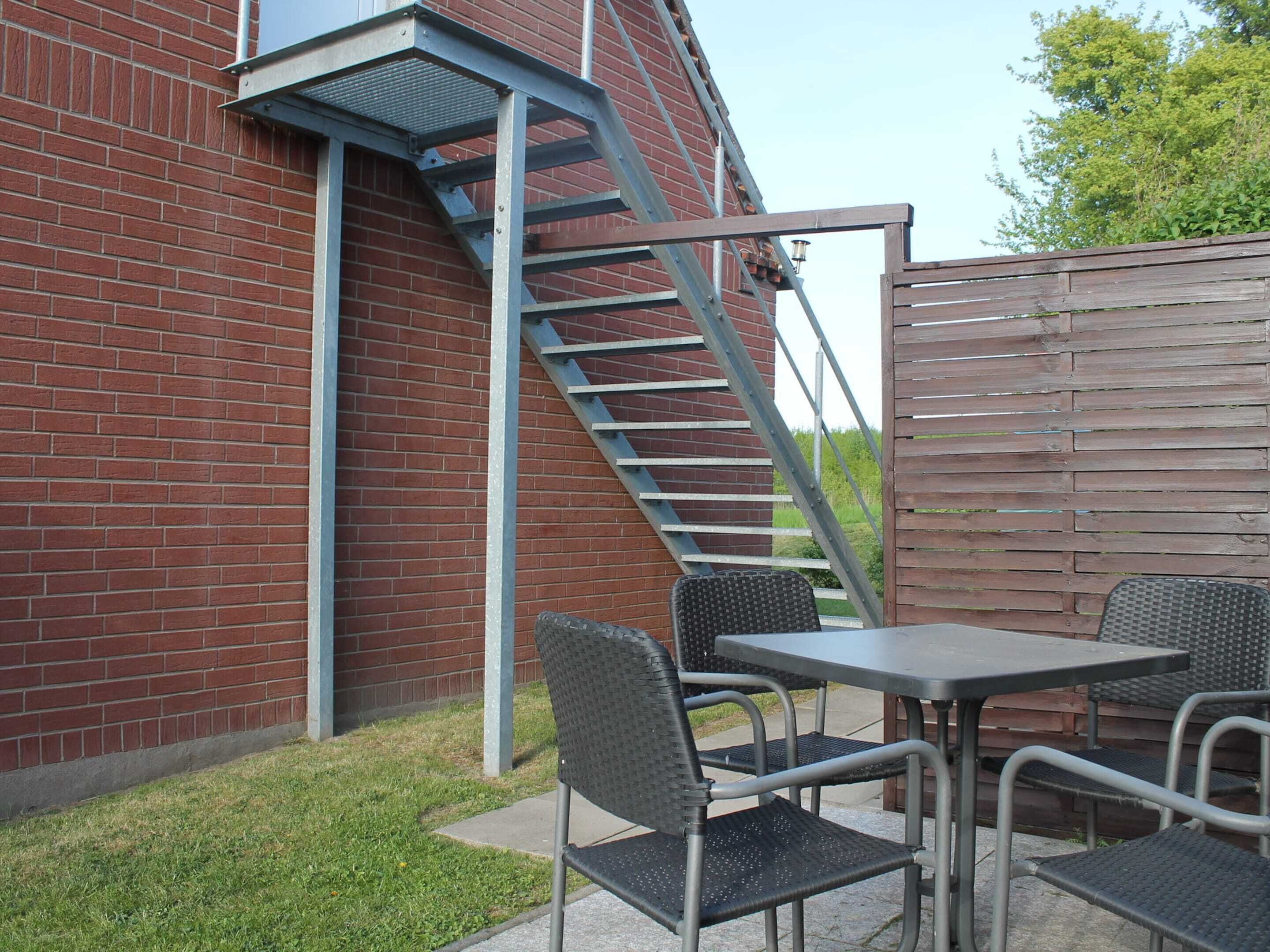 A brick building with a metal staircase, outdoor seating with metal chairs and a table, and a wooden fence.