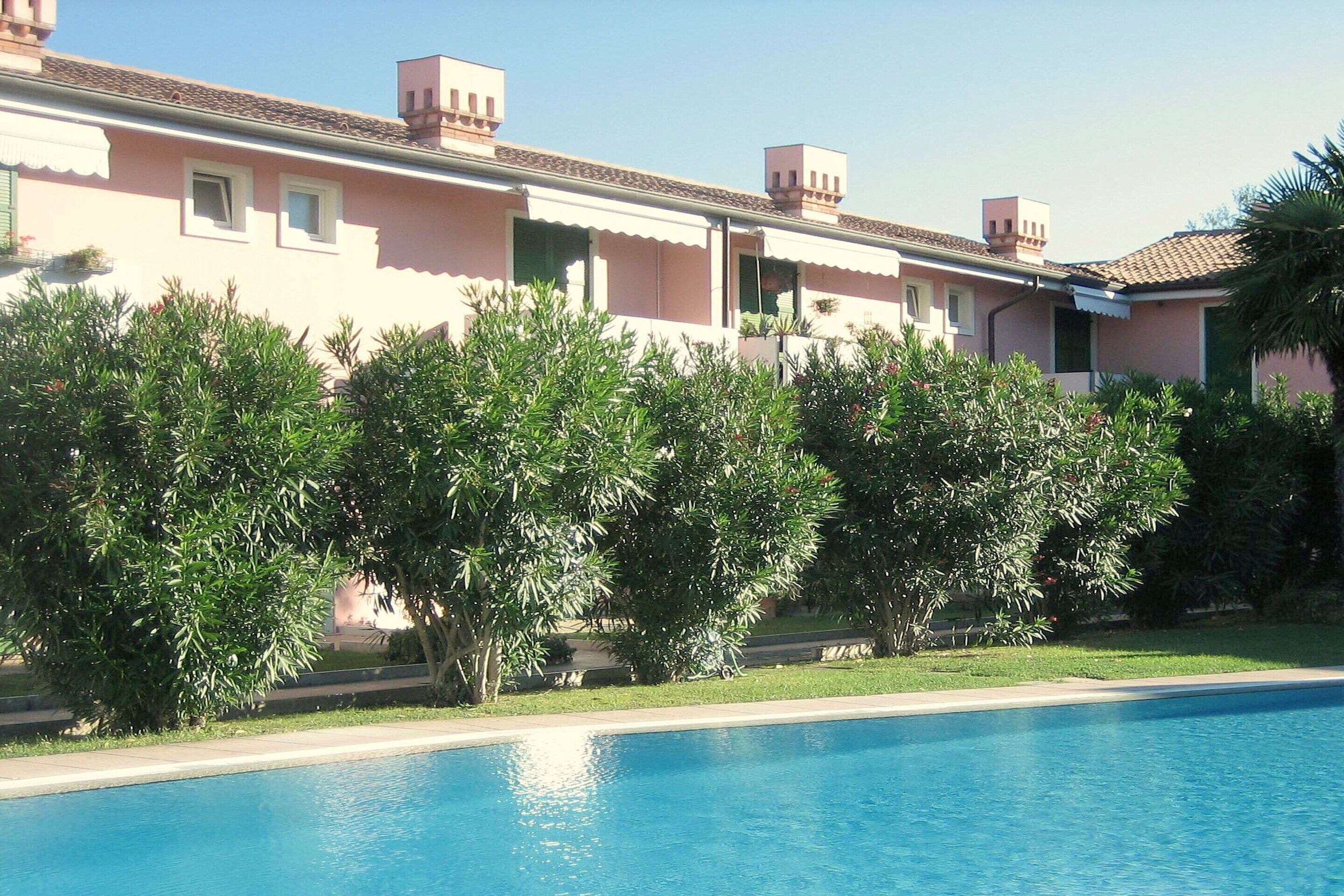 A swimming pool with a clear blue water surface, surrounded by lush green trees and a pink building with multiple windows and balconies.