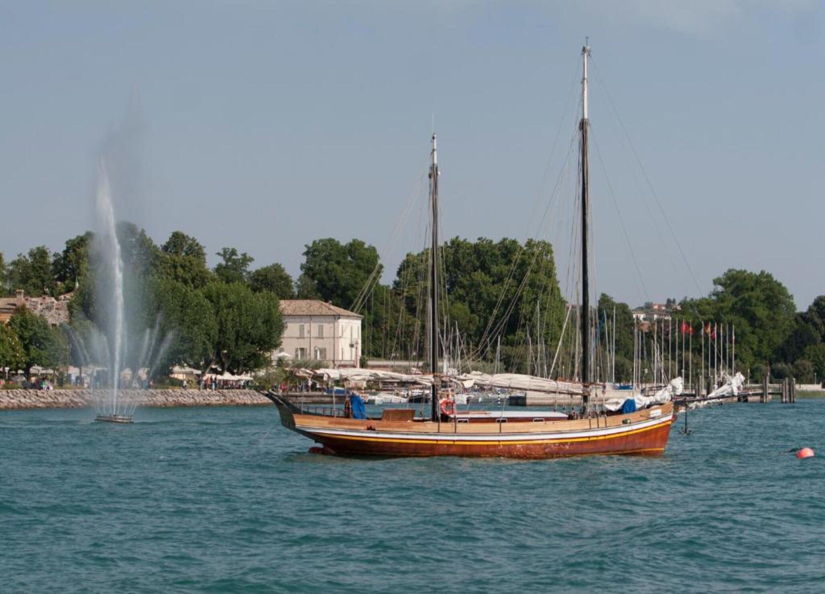 Un bateau en bois traditionnel est amarré près d’une fontaine, avec en arrière-plan des arbres et un bâtiment.
