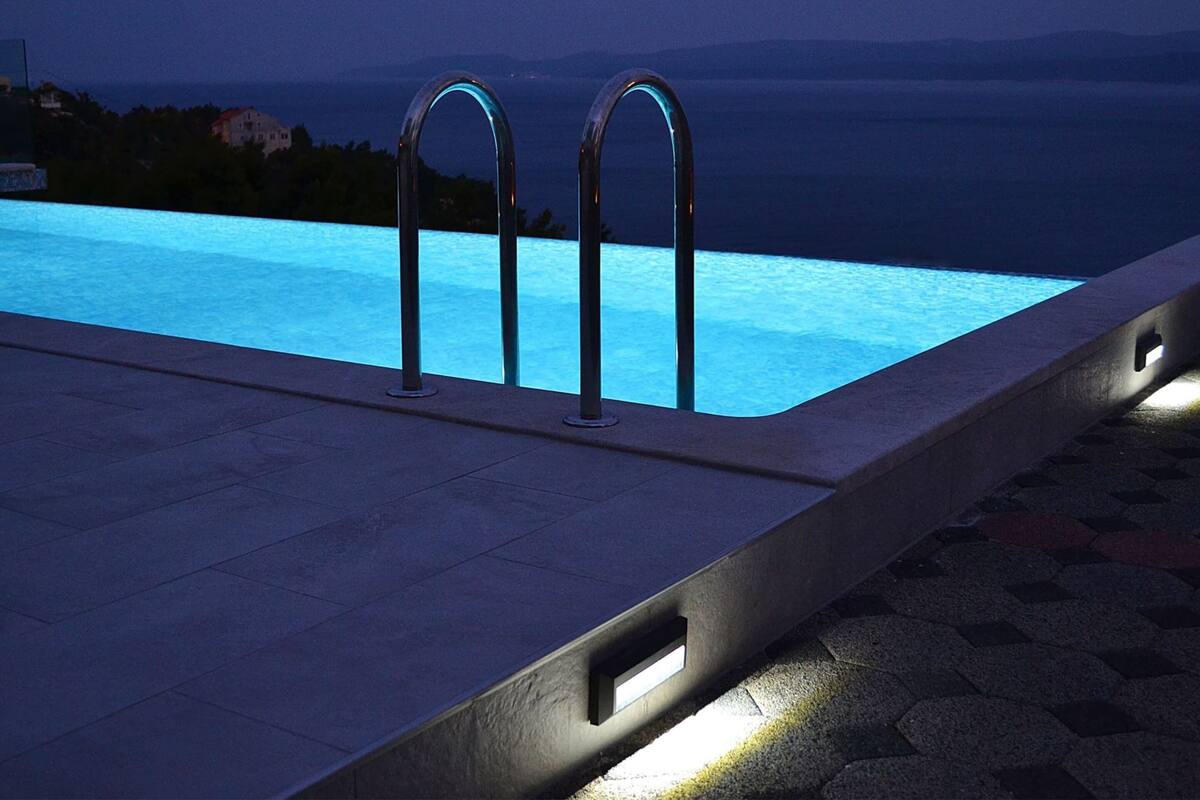 A swimming pool with two metal handrails, a view of the sea, and a tiled deck.
