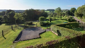 A rural landscape with a fenced area, a paved path, and a variety of trees and shrubs. - Cottage in a Well-equipped and Comfortable Farmhouse (Marche-en-Famenne)