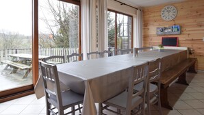 A dining area with a long table, chairs, and a bench. There is a large window with curtains, a wooden wall with a clock, and a small shelf with books. - Cottage in a Well-equipped and Comfortable Farmhouse (Marche-en-Famenne)