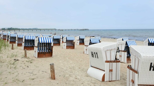 Strandstühle mit gestreiften Sonnenschirmen, angeordnet auf dem Sand, ein Holzsteg im Hintergrund und ein klarer Himmel.