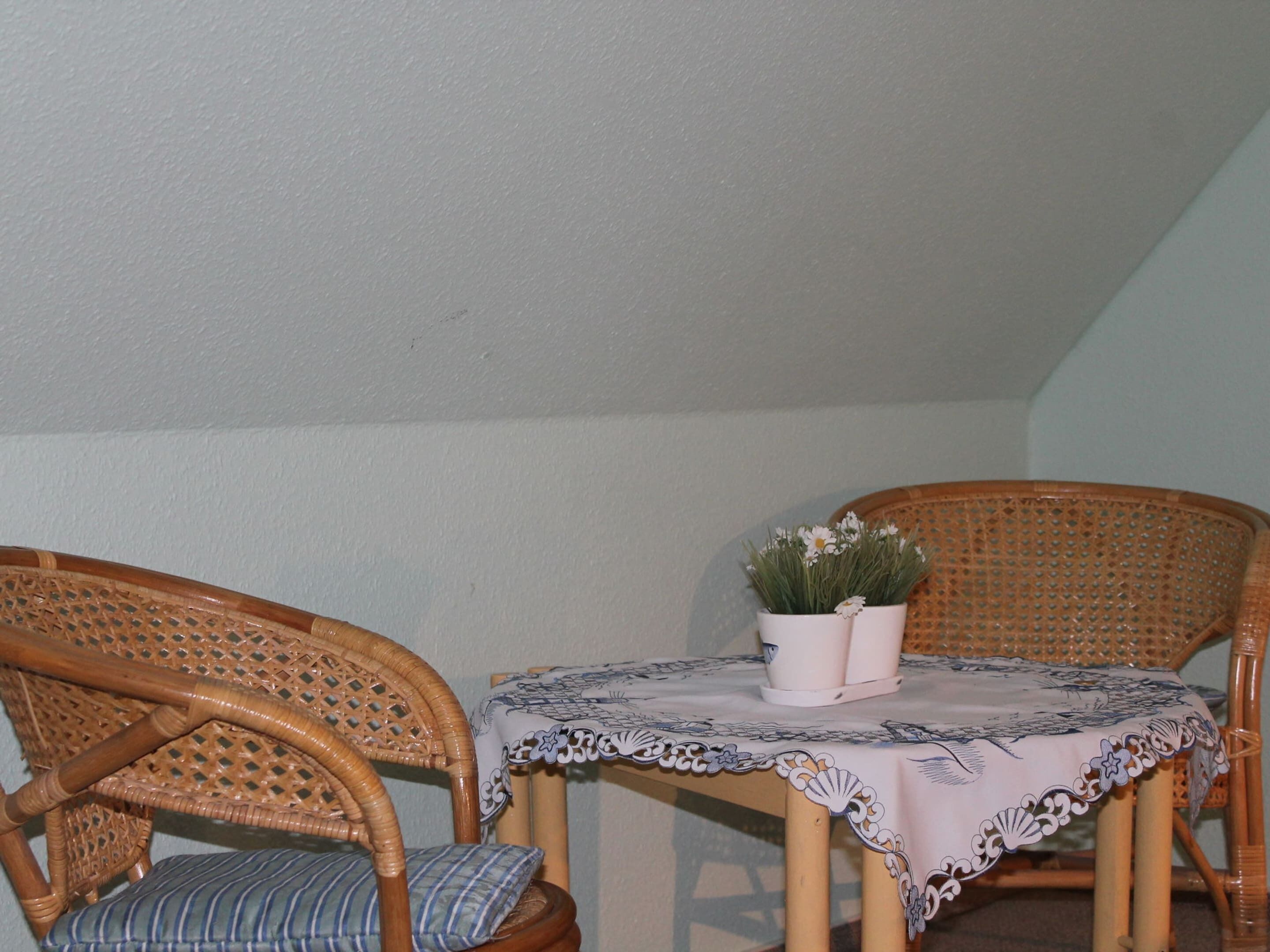 Une petite table ronde recouverte d’une nappe en dentelle, deux chaises en osier et un pot de fleurs orné de fleurs blanches.